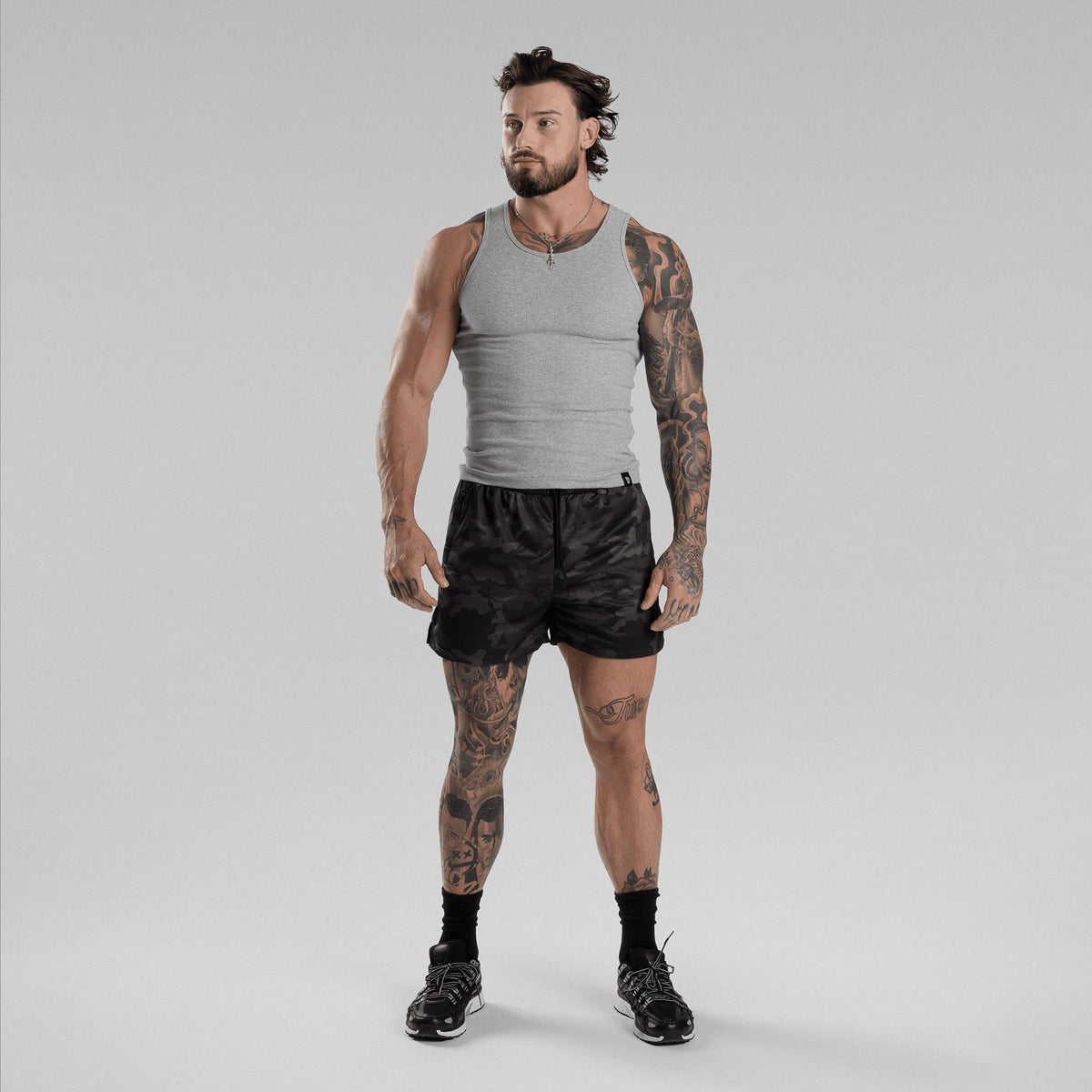 A man wearing a gray ribbed tank top, black camouflage athletic shorts, black socks, and black sneakers.